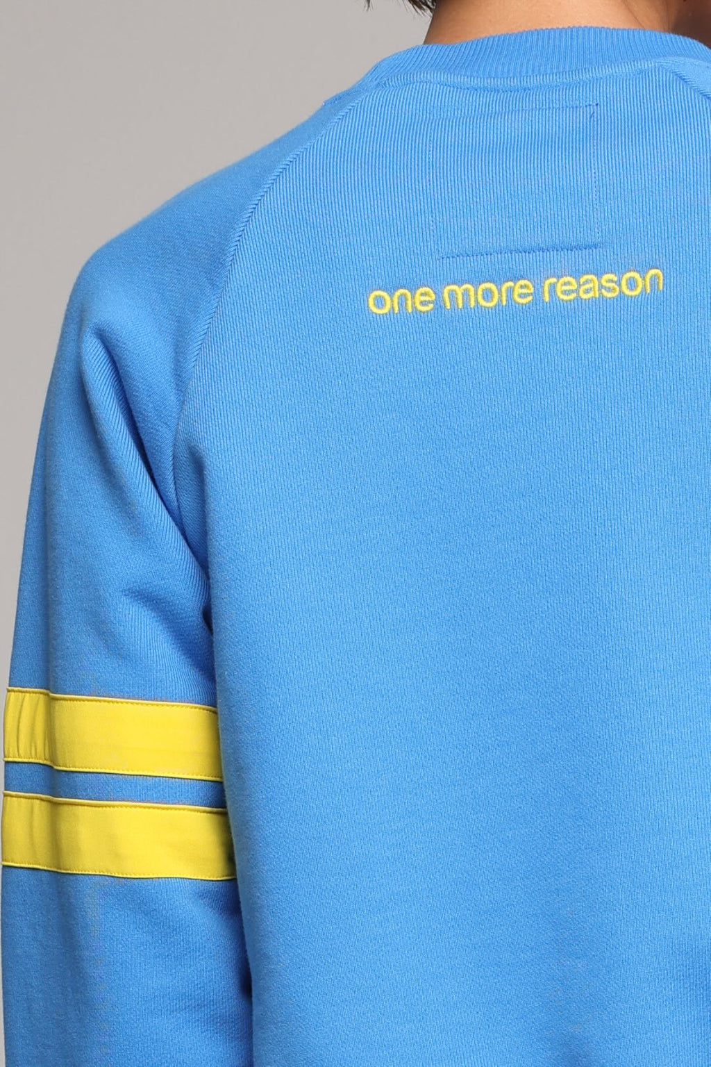 Close-up back view of a person wearing a bright blue sweatshirt with yellow text reading 'one more reason' across the upper back and two horizontal yellow stripes on the sleeve.