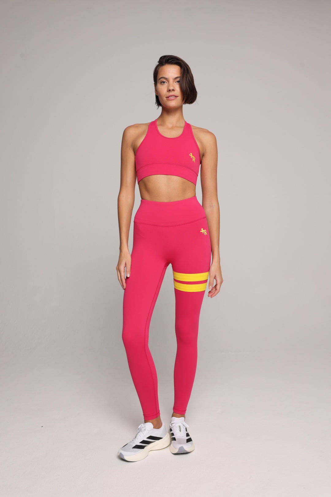 A woman wearing a bright pink performance tank top and leggings with yellow stripes by One More Reason, the premium sportswear brand made in Europe in a studio setting 