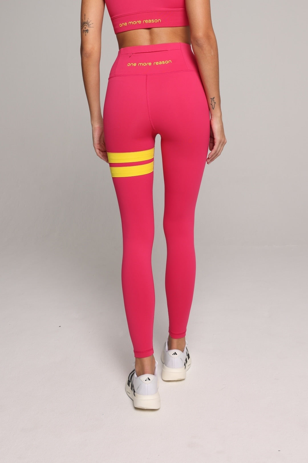 A woman seen from the back wearing bright pink performance leggings with yellow stripes by One More Reason, the premium sportswear brand made in Europe in a studio setting 