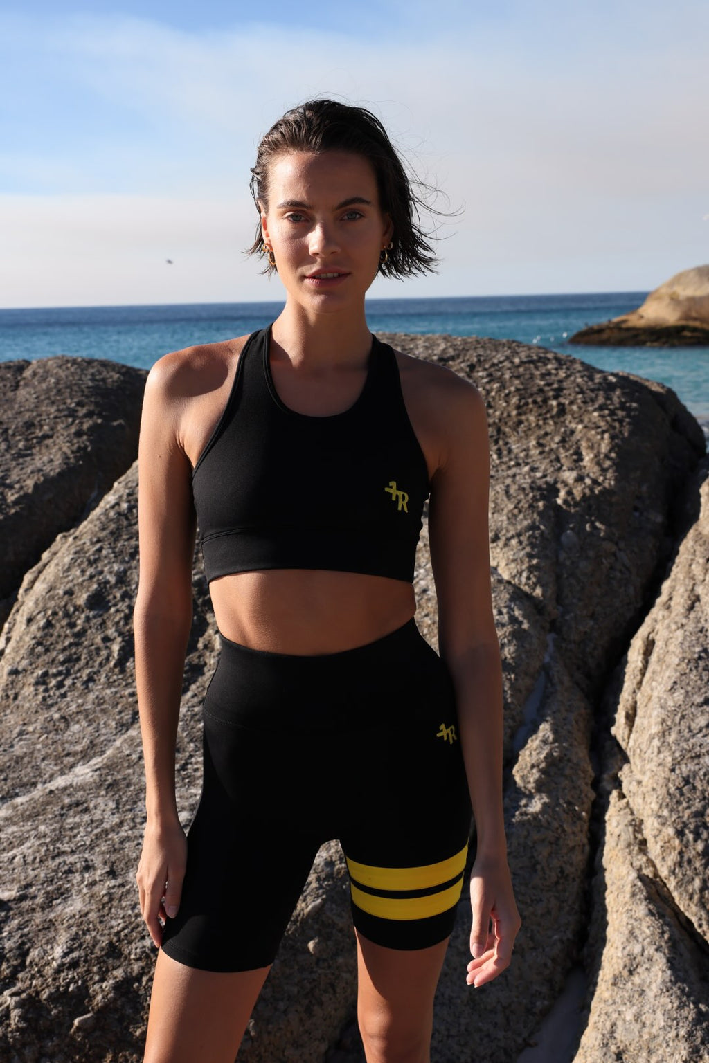 Woman in athletic black wear with yellow stripes by one more reason standing on rocks by the ocean