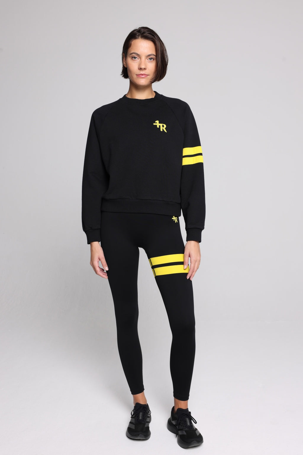 Woman wearing a black crewneck sweatshirt with yellow stripes on the sleeve and a yellow embroidered logo on the chest, paired with black leggings featuring yellow stripes on the thigh. All from One More Reason, the European Premium Sportswear Brand.