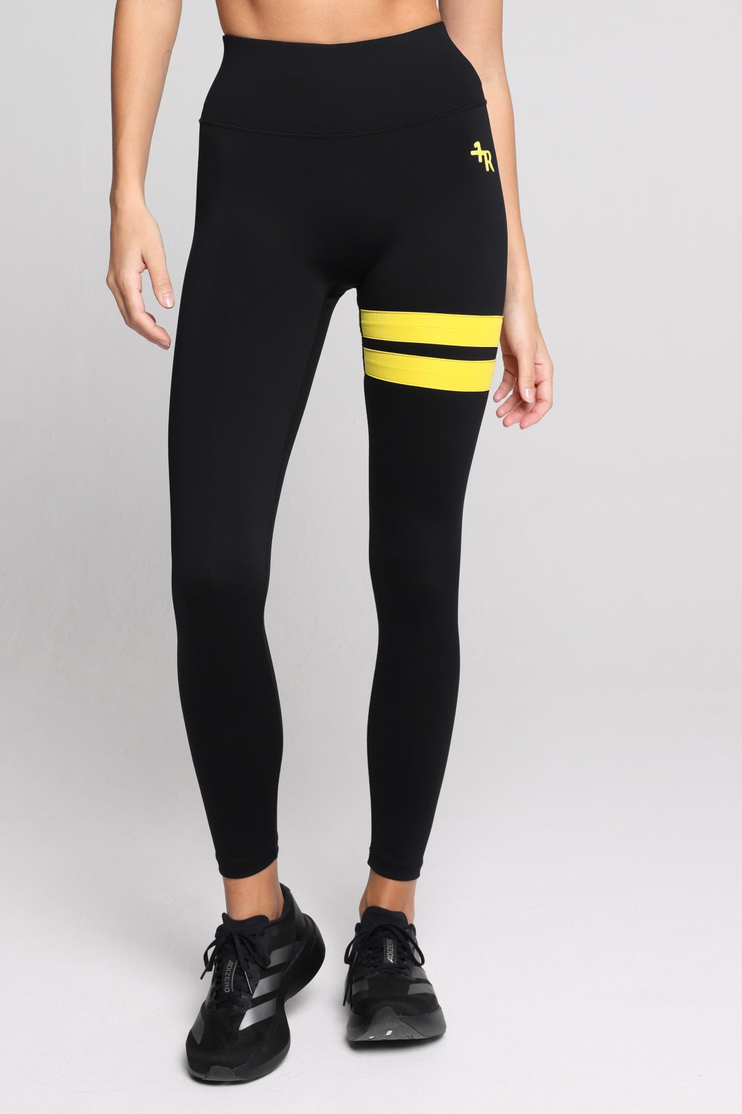 Black high-waisted leggings with two horizontal yellow stripes on the upper right thigh and a small yellow logo on the left hip, shown on a model wearing black sneakers against a light gray studio background. All by One More Reason.