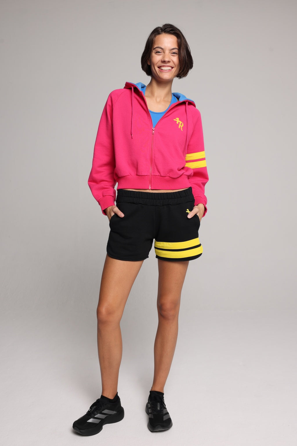 A woman wearing a bright pink zip-up hoodie with a blue hood lining, yellow graphic logo on the chest, and yellow stripes on the sleeve, paired with black shorts featuring yellow stripes, against a solid gray background. All from One More Reason, the European Premium Sportswear Brand.