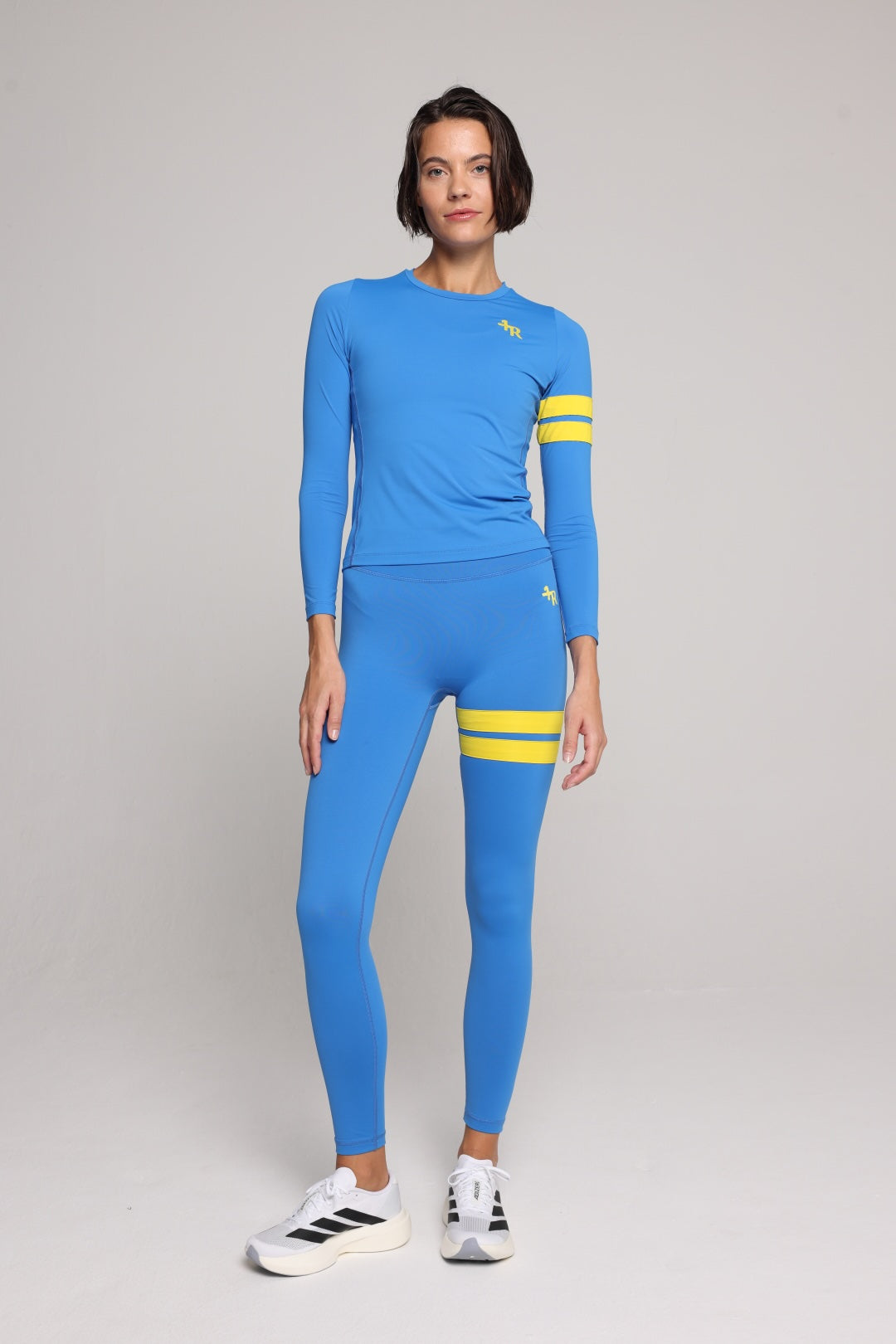 A woman in a bright blue long-sleeve athletic top and matching leggings set with yellow stripe accents on the sleeves and thighs, against a plain light gray studio background. All by One More Reason, the European premium sportswear brand.