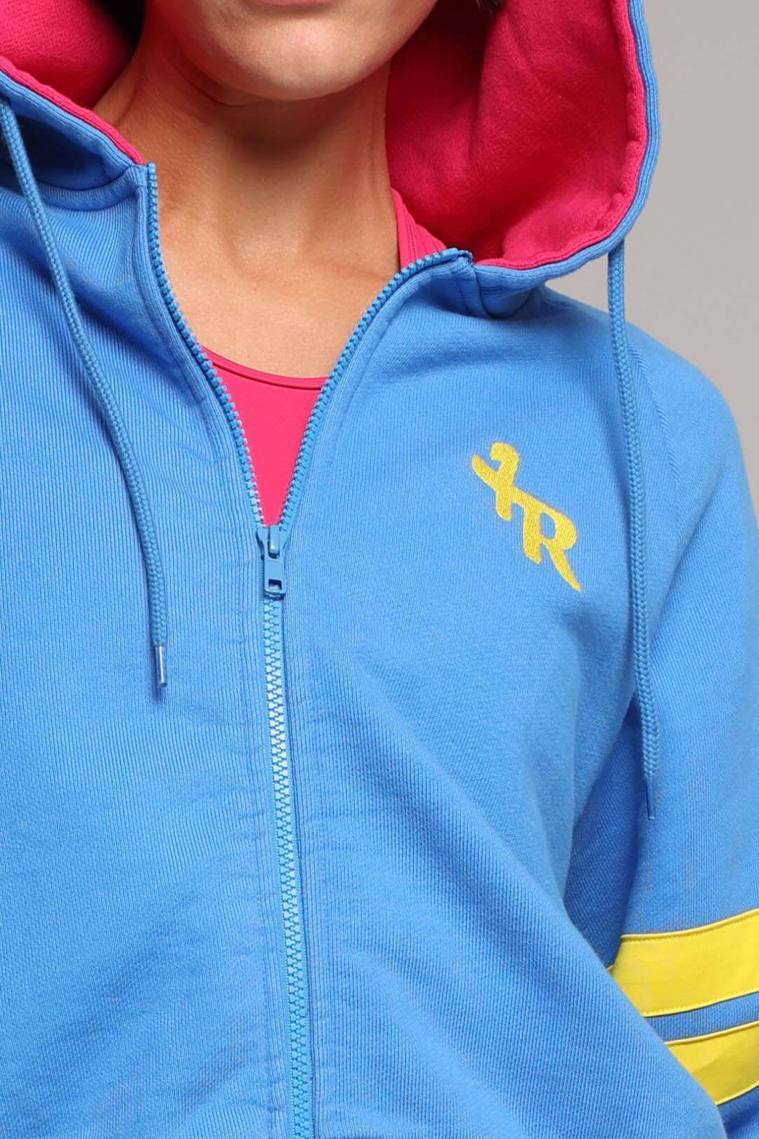 A close-up of a woman wearing a bright blue zip-up hoodie with a pink-lined hood, a yellow embroidered monogram on the chest, and yellow stripes on the sleeve from One More Reason, the European Premium Sportswear Brand.
