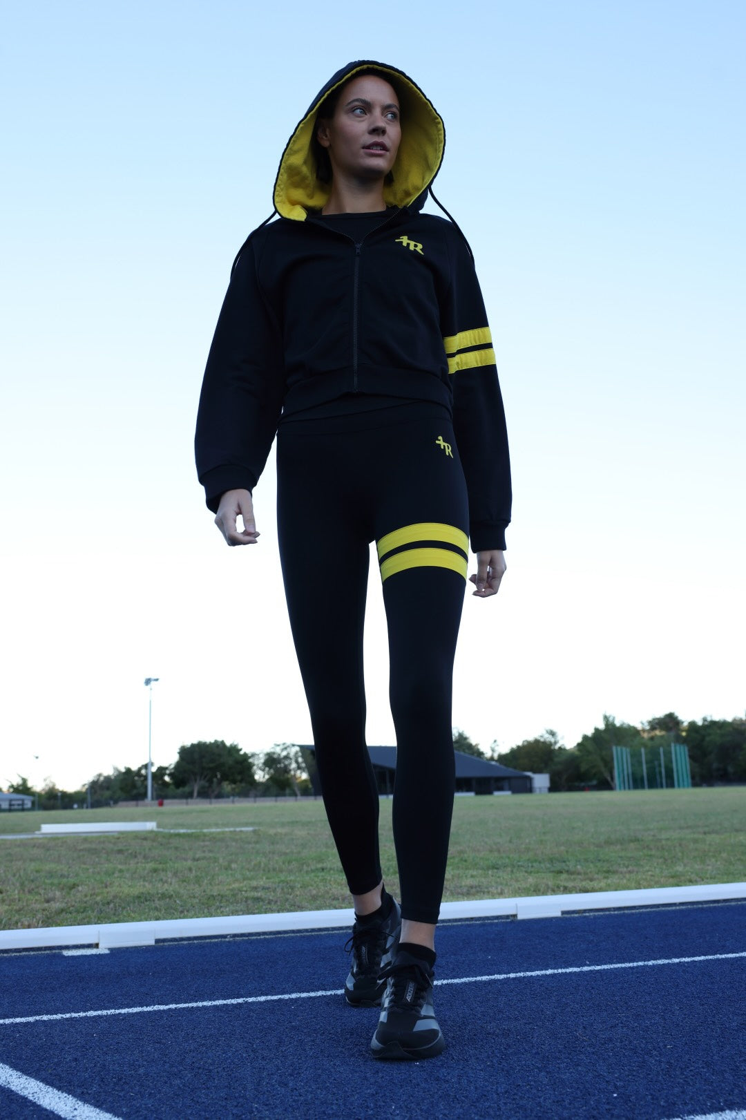 A woman wearing a black zip-up hoodie and matching black leggings with bright yellow accents, photographed outdoors against a clear blue sky. All from One More Reason, the European Premium Sportswear Brand.