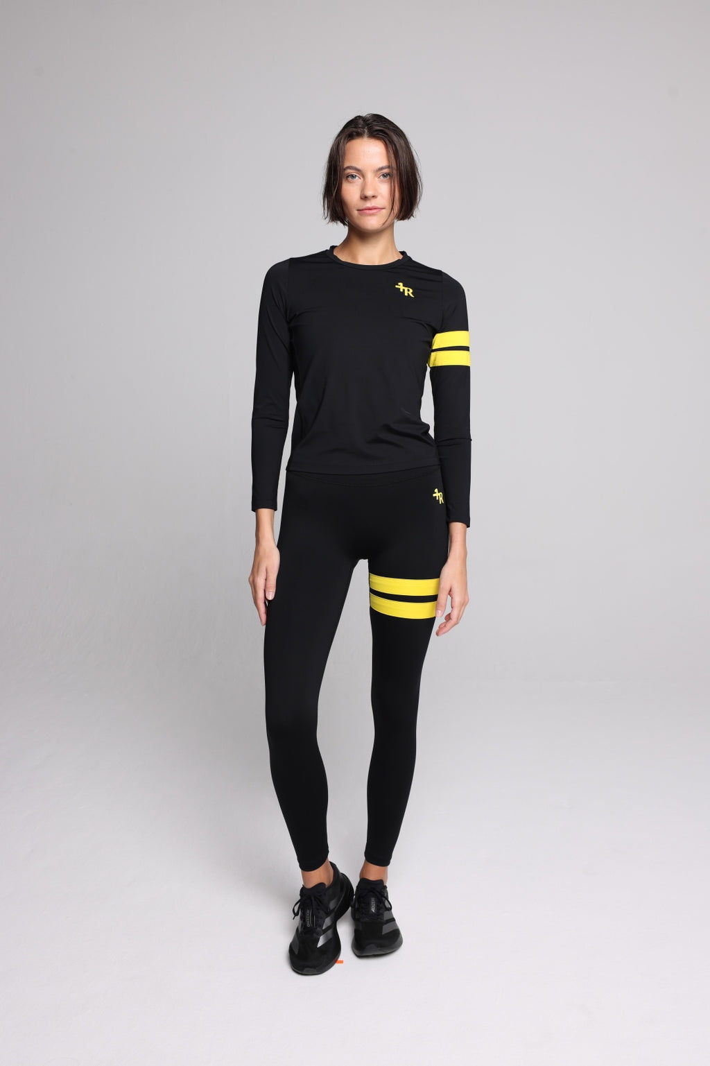 A woman wearing a black long-sleeve athletic top and matching black leggings with yellow stripe accents on the sleeves and thighs, standing against a light gray studio background. Sportswear by one more reason, the premium sportswear brand, crafted in Europe.