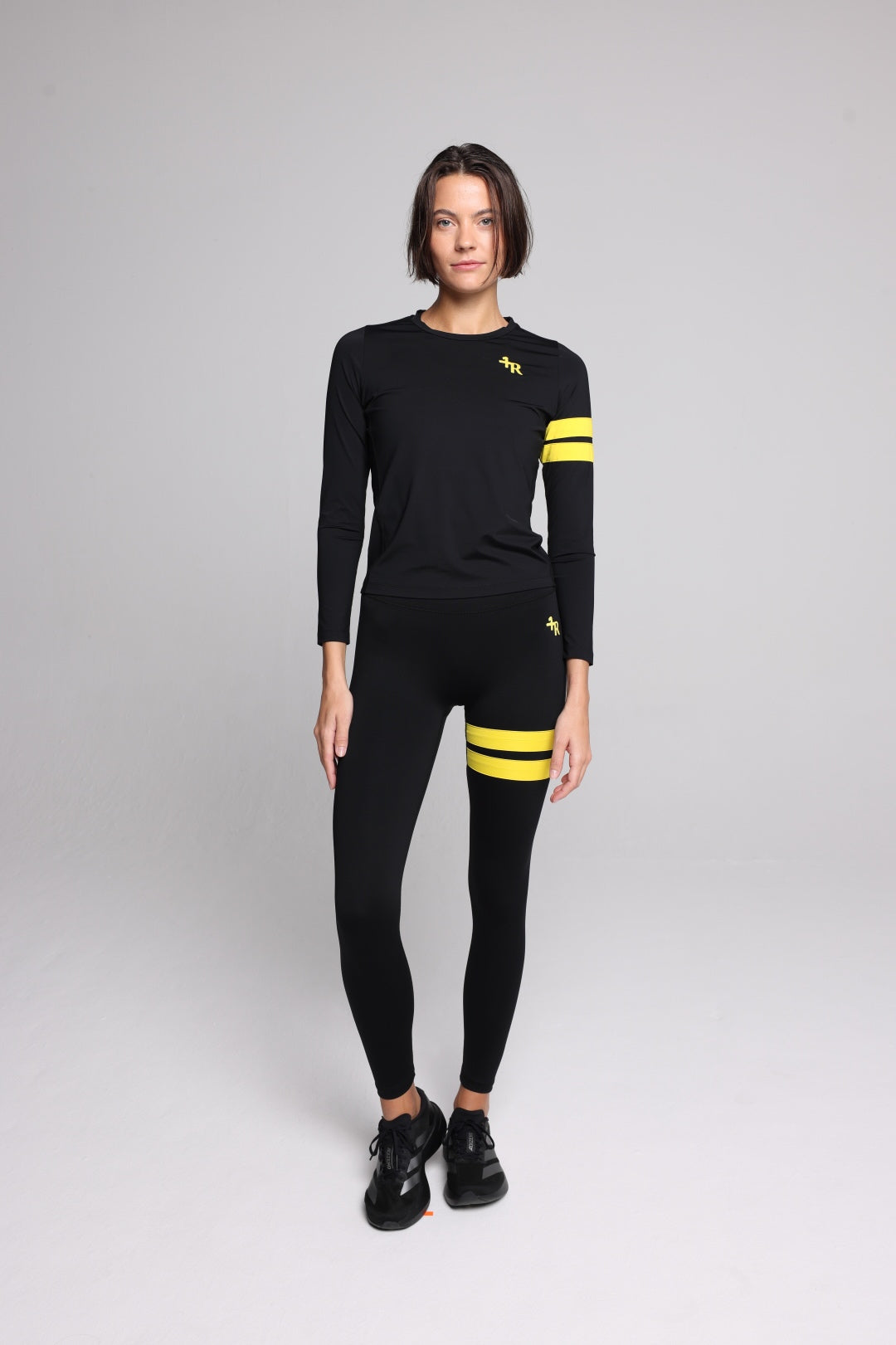 A woman wearing a black long-sleeve athletic top and matching black leggings with yellow stripe accents on the sleeves and thighs, standing against a light gray studio background. Sportswear by one more reason, the premium sportswear brand, crafted in Europe.