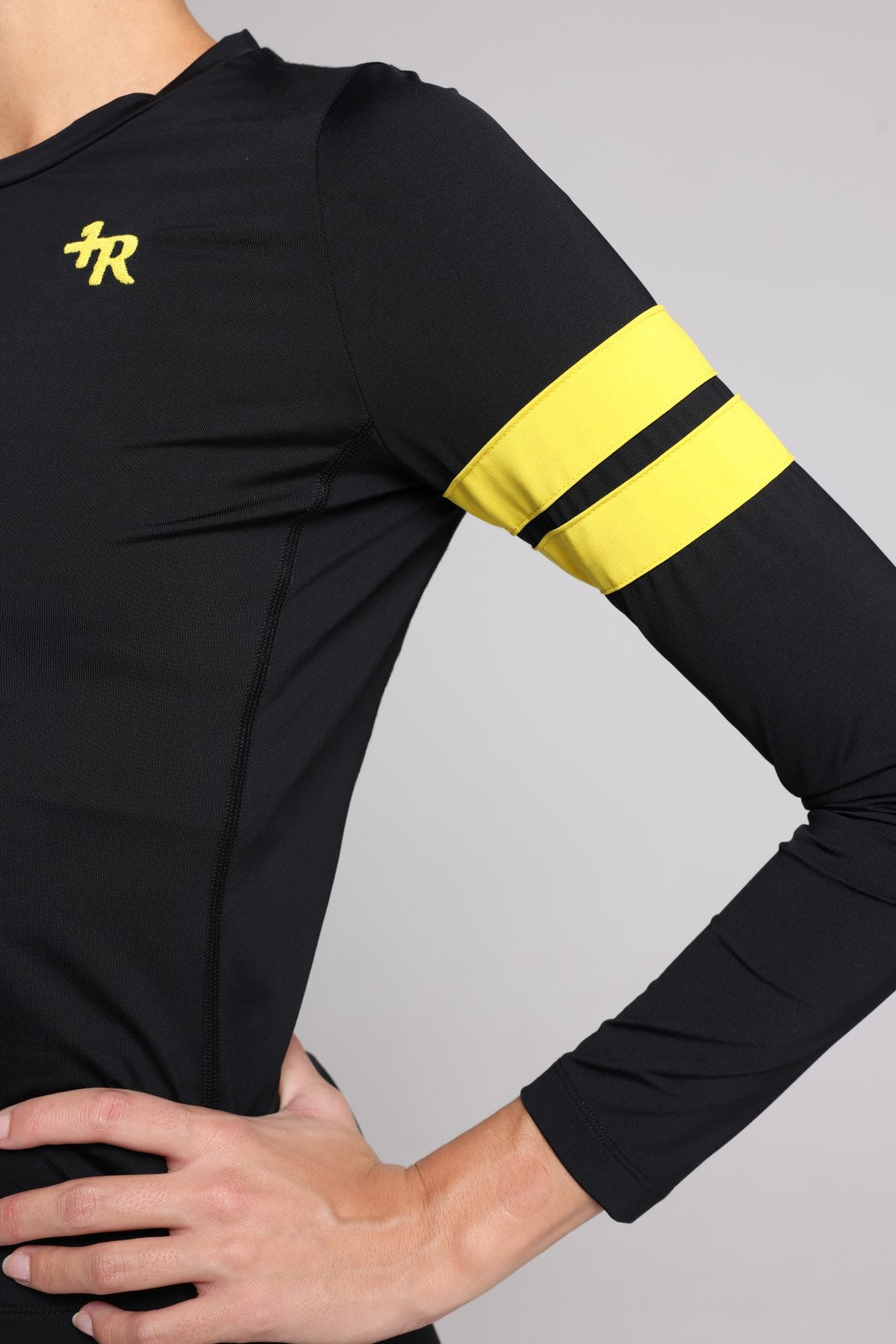A close-up of a black long-sleeve athletic top worn by a model, featuring a yellow stylized logo on the left chest and two horizontal yellow stripes on the right sleeve, set against a solid gray background.