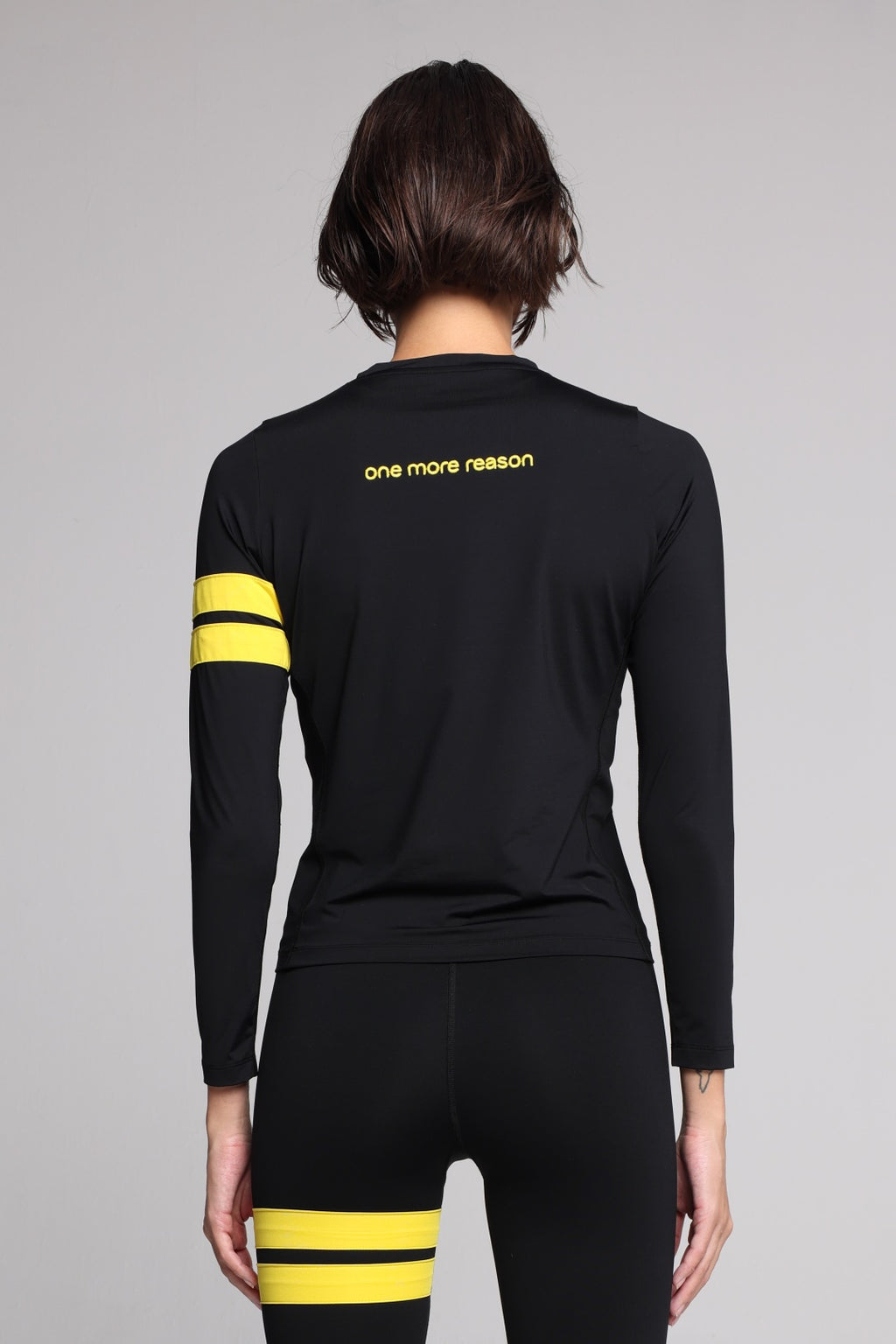A woman shown from the back wearing a black long-sleeve athletic top with yellow stripes on the sleeves and yellow text reading 'one more reason' on the upper back, paired with black leggings with yellow stripes near the upper thigh, against a light gray studio background.
