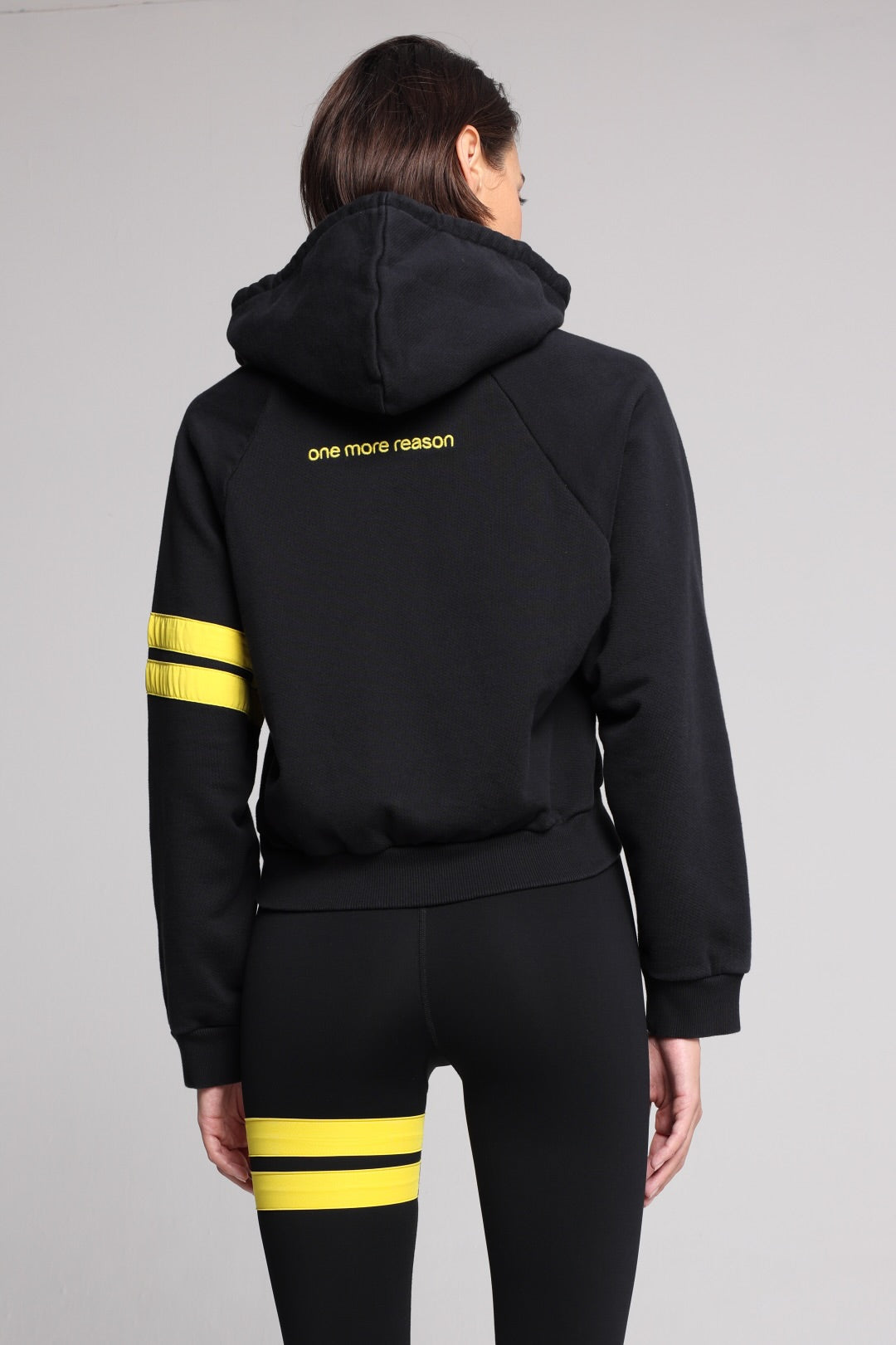 A woman shown from the back wearing a black hooded sweatshirt and matching black leggings, both featuring yellow stripe accents on the sleeves and thighs. All from One More Reason, the European Premium Sportswear Brand.