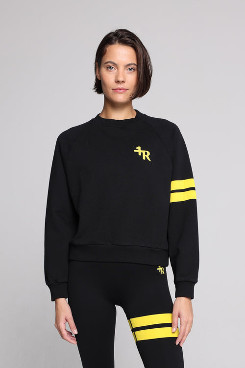 Woman wearing a black crewneck sweatshirt with yellow stripes on the sleeve and a yellow embroidered logo on the chest, paired with black leggings featuring yellow stripes on the thigh. All from One More Reason, the European Premium Sportswear Brand.