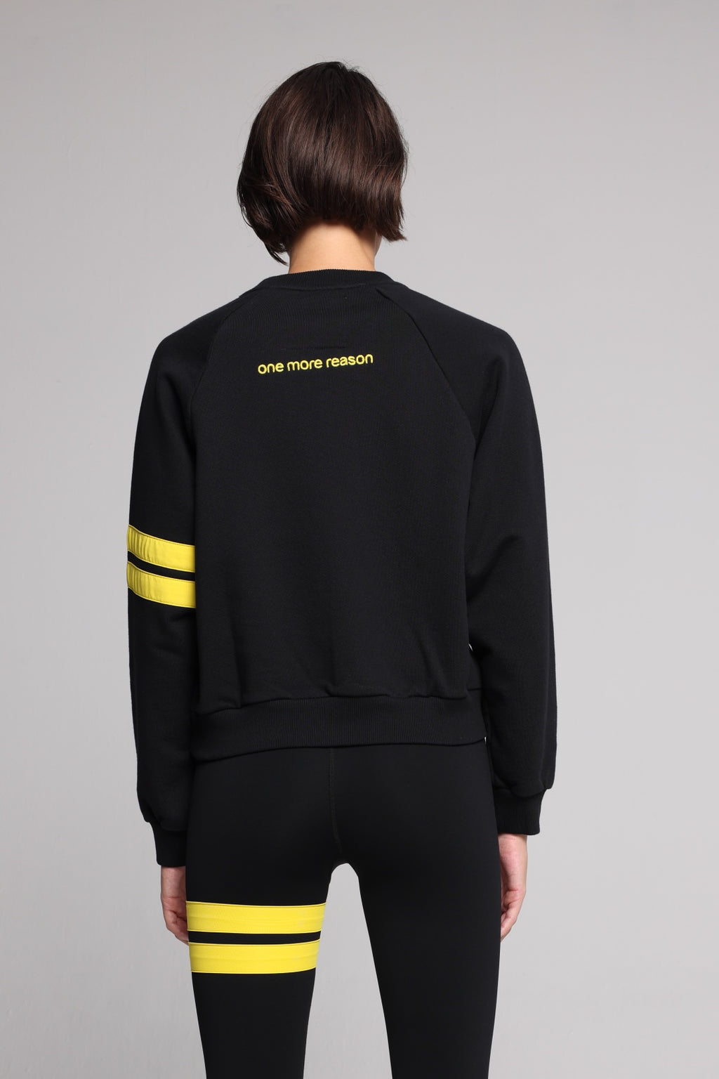 A woman showing from the back wearing a black crewneck sweatshirt with yellow stripes on the sleeve and a yellow embroidered logo on the back, paired with black leggings featuring yellow stripes on the thigh. All from One More Reason, the European Premium Sportswear Brand.