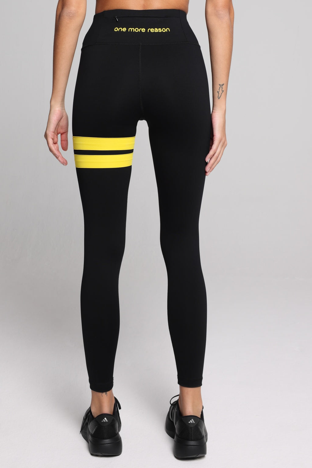 Black high-waisted leggings with two horizontal yellow stripes on the upper thigh and yellow text on the waistband reading 'one more reason', shown on a model from the back against a light gray studio background. All by One More Reason.