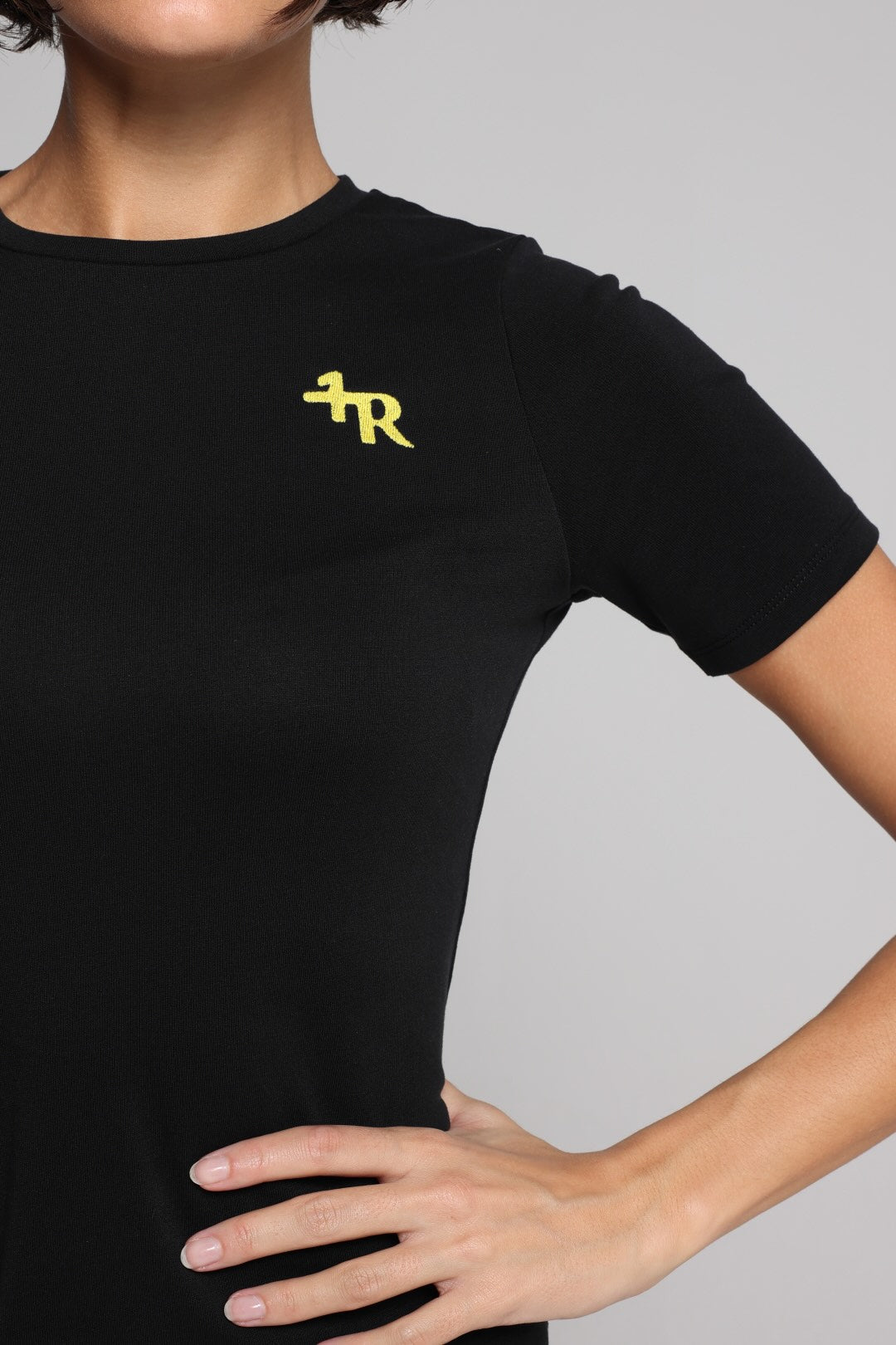 A close-up of a woman wearing a black short-sleeve crewneck t-shirt with a small yellow stylized logo on the left chest, set against a solid gray background.
