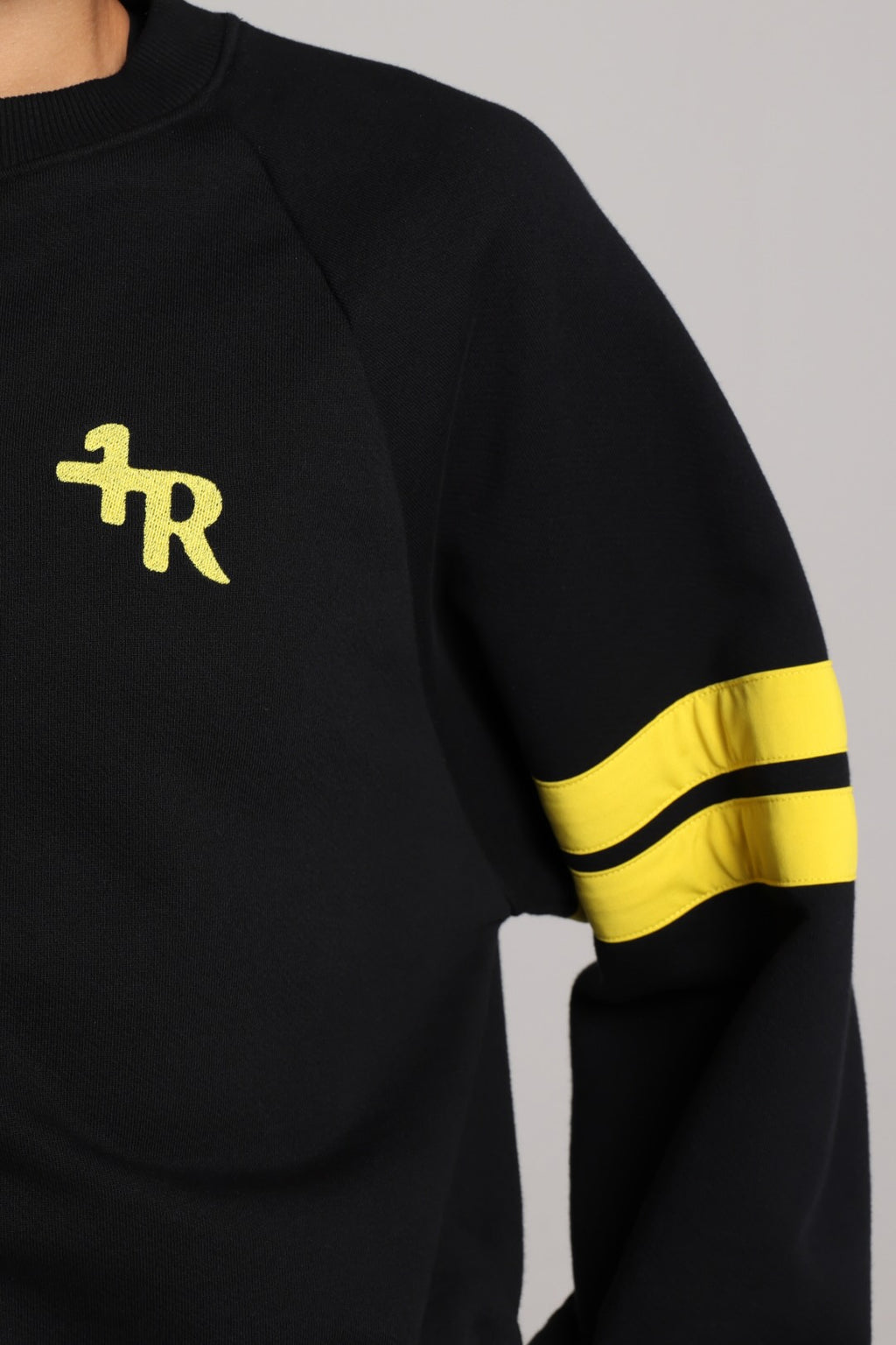 Close-up of a black long-sleeve sweatshirt with a yellow embroidered logo on the chest and two yellow stripes on the sleeve. All from One More Reason, the European Premium Sportswear Brand.