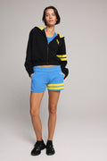 A woman wearing a black cropped zip-up hoodie with yellow stripes, a blue crop top, bright blue shorts with yellow stripes, and black sneakers. All from One More Reason, the European premium sportswear brand.