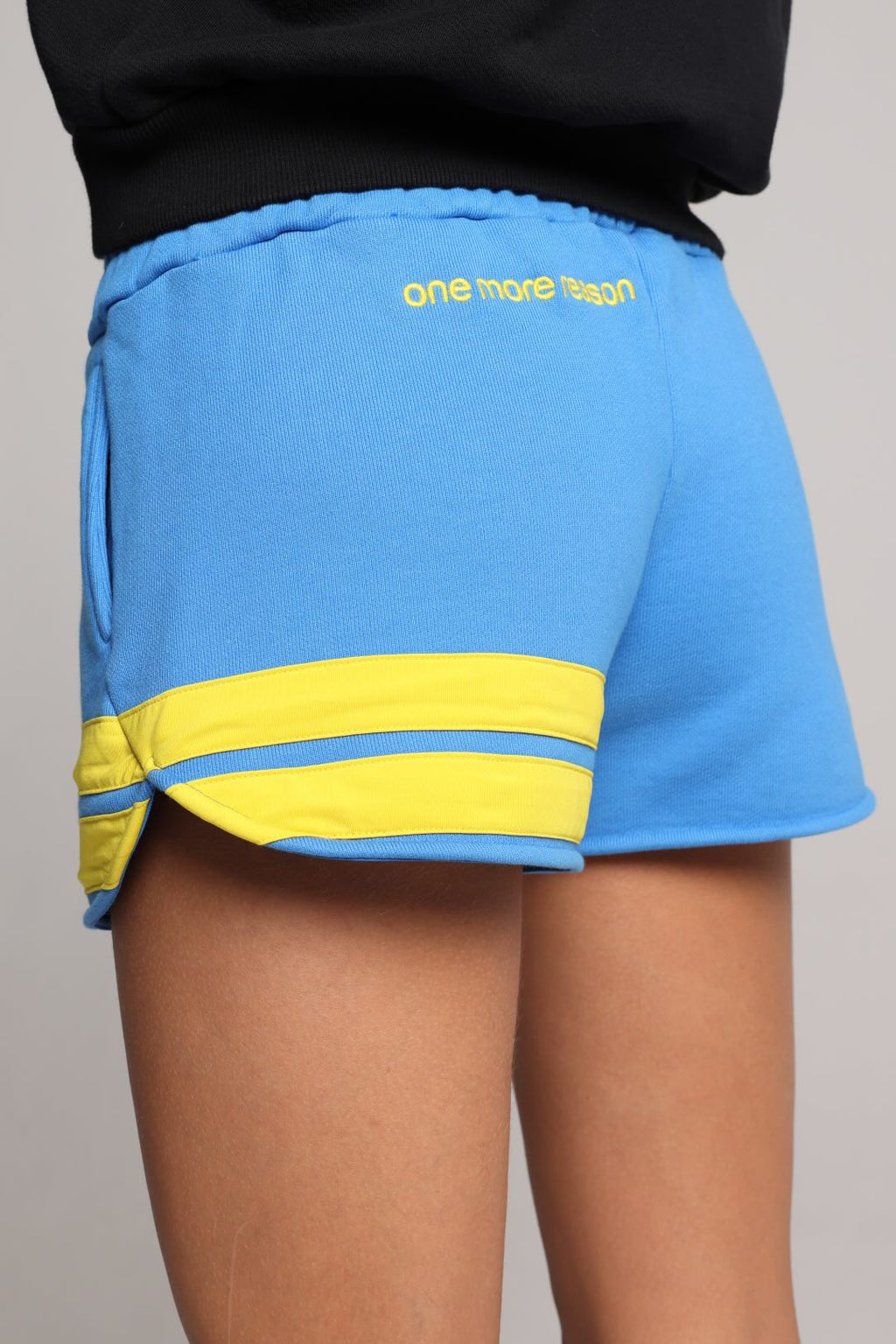 A close-up of a person wearing bright blue athletic shorts with a yellow stripe detail and yellow embroidered text reading 'one more try' on the waistband, paired with a black top against a plain light gray background.