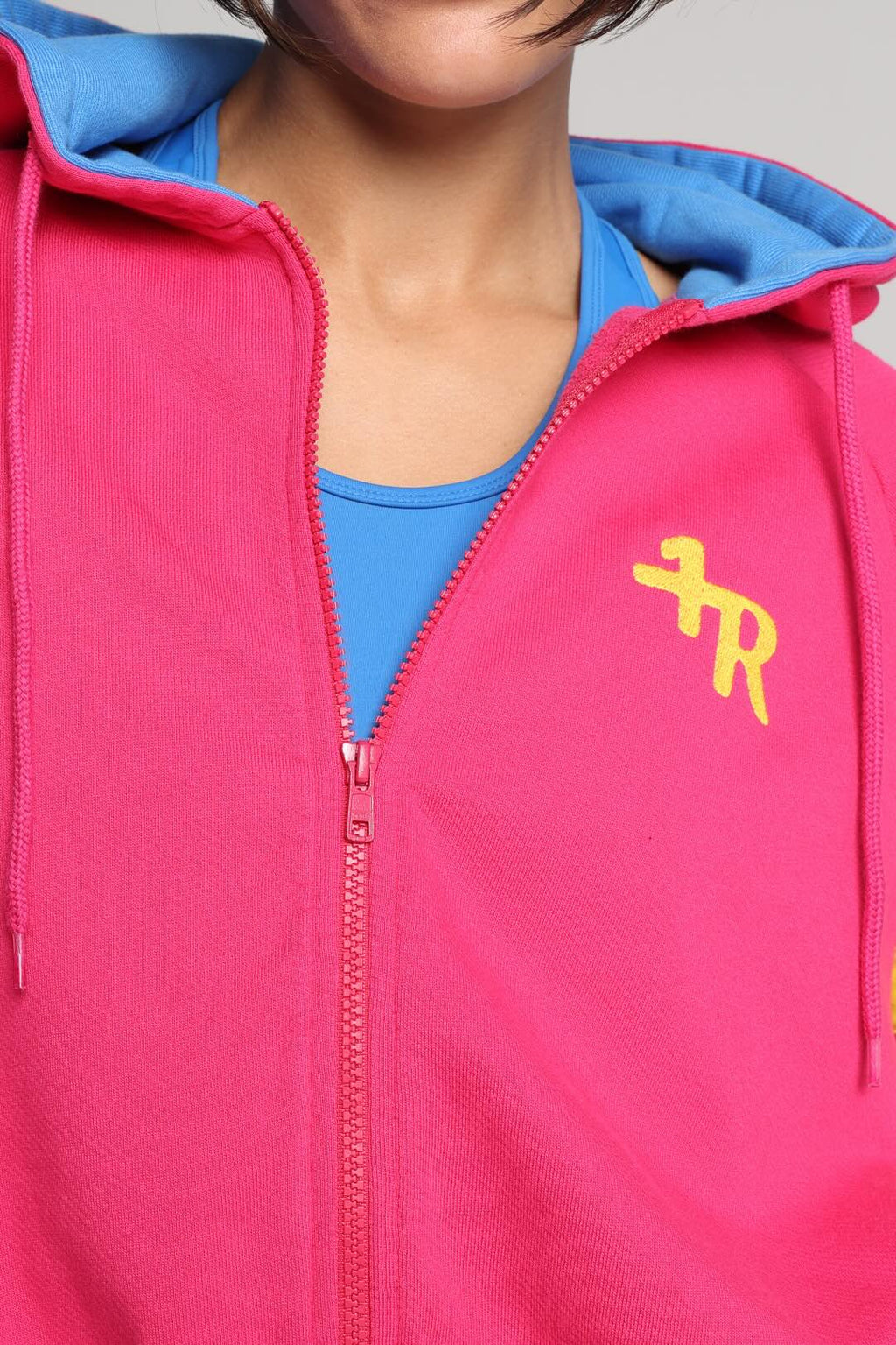 A close-up of a woman wearing a bright pink zip-up hoodie with a blue hood lining and a yellow stylized ogo on the left chest, layered over a blue top. All from One More Reason, the European Premium Sportswear Brand.