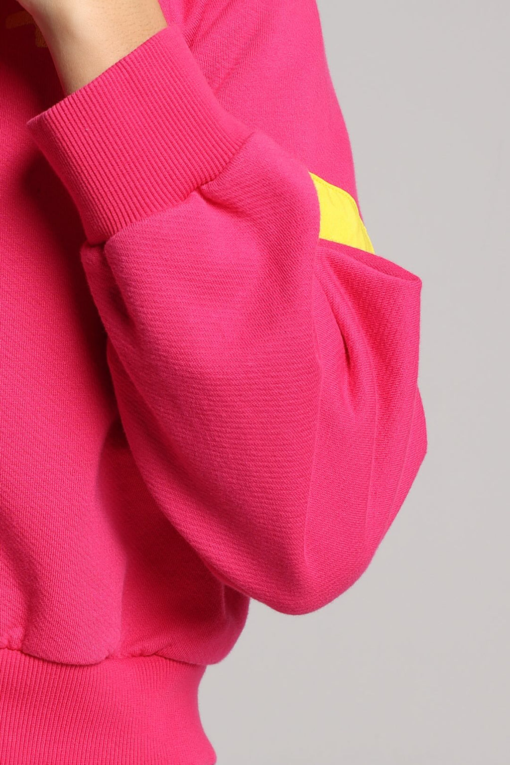 A close-up of a bright pink ribbed sweatshirt sleeve with a small yellow geometric patch on the upper cuff, shown against a light gray background.