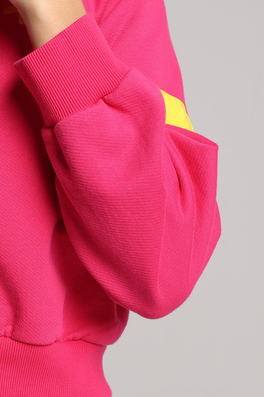 A close-up of a bright pink ribbed sweatshirt sleeve with a small yellow geometric patch on the upper cuff, shown against a light gray background.