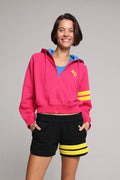 A woman wearing a bright pink zip-up hoodie with a blue hood lining, yellow graphic logo on the chest, and yellow stripes on the sleeve, paired with black shorts featuring yellow stripes, against a solid gray background. All from One More Reason, the European Premium Sportswear Brand.