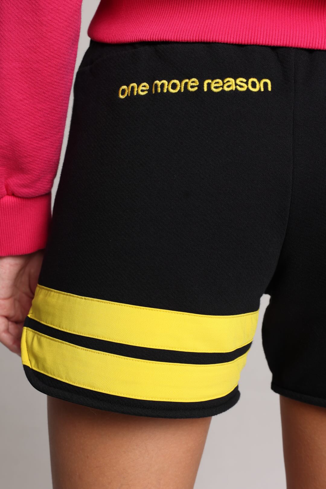 Black athletic shorts with yellow stripes and 'one more reason' text on the back, worn by a model in a pink top.
