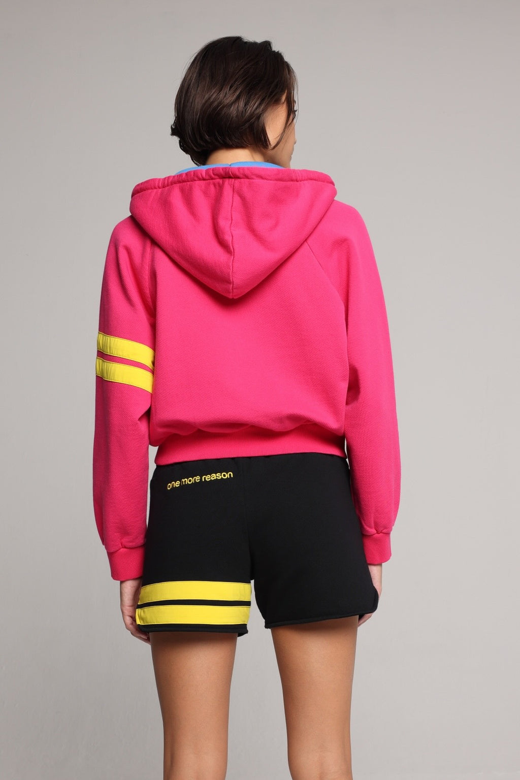 woman wearing a premium cotton pink hoodie with a blue hood and black shorts with yellow stripes by one more reason back view studio set up