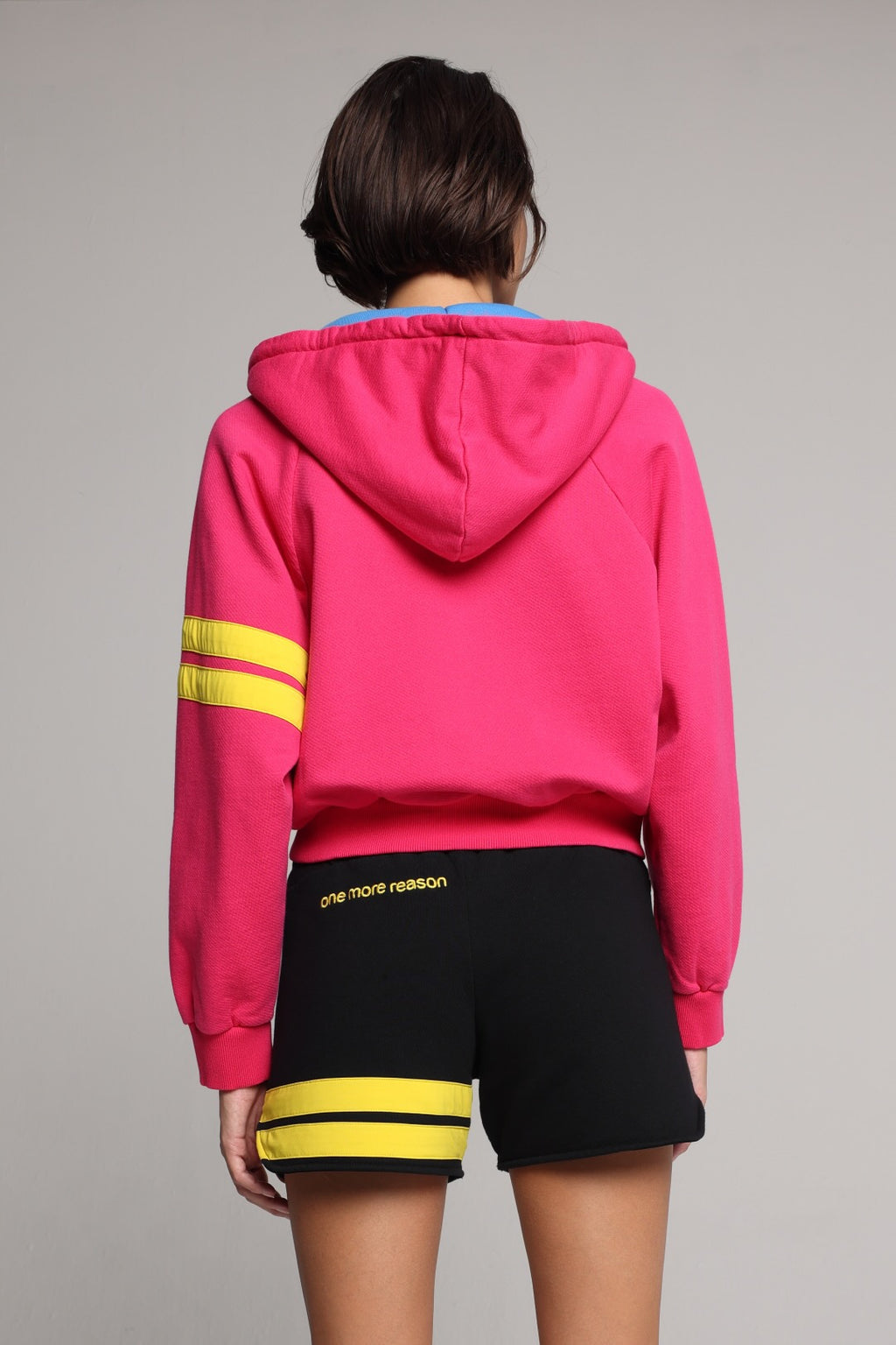 A woman showing from the back wearing a bright pink zip-up hoodie with a blue hood lining, and yellow stripes on the sleeve, paired with black shorts featuring yellow stripes. All from One More Reason, the European Premium Sportswear Brand.
