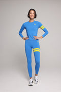 A woman modeling a bright blue long-sleeve athletic top and matching leggings set with yellow stripe accents on the sleeves and upper thighs, photographed against a neutral light gray studio background.
