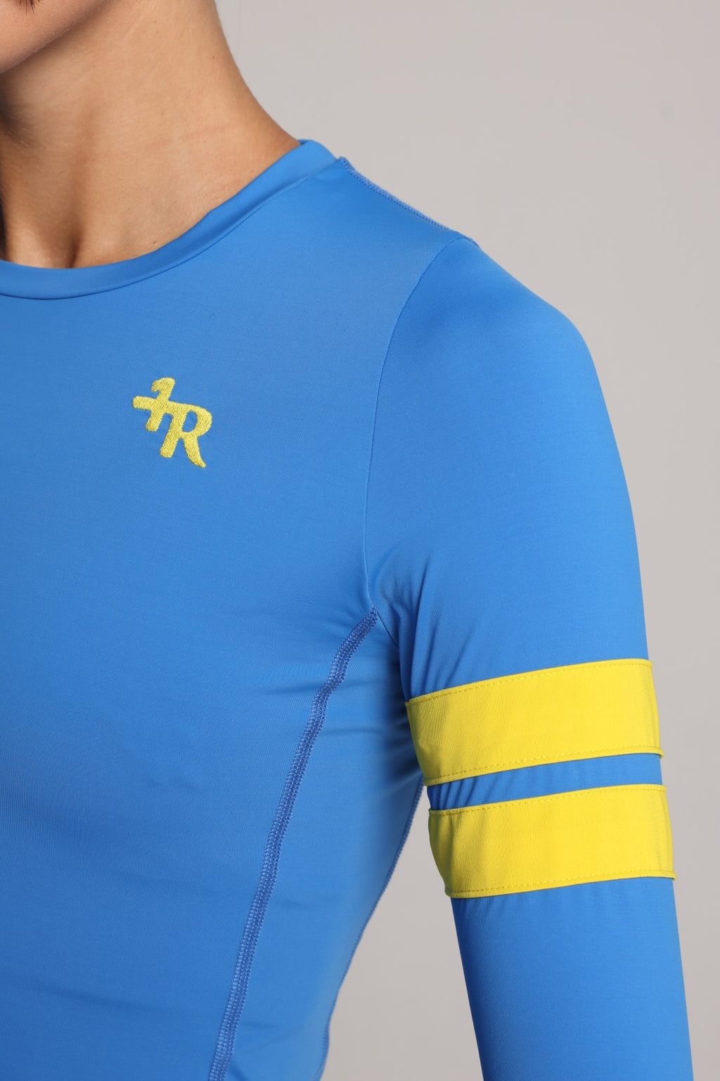 Close-up of a blue long-sleeve athletic top worn by a model, featuring a yellow stylized logo on the left chest and two horizontal yellow stripes on the right sleeve, against a light gray background - by One More Reason, the European premium sportswear brand.