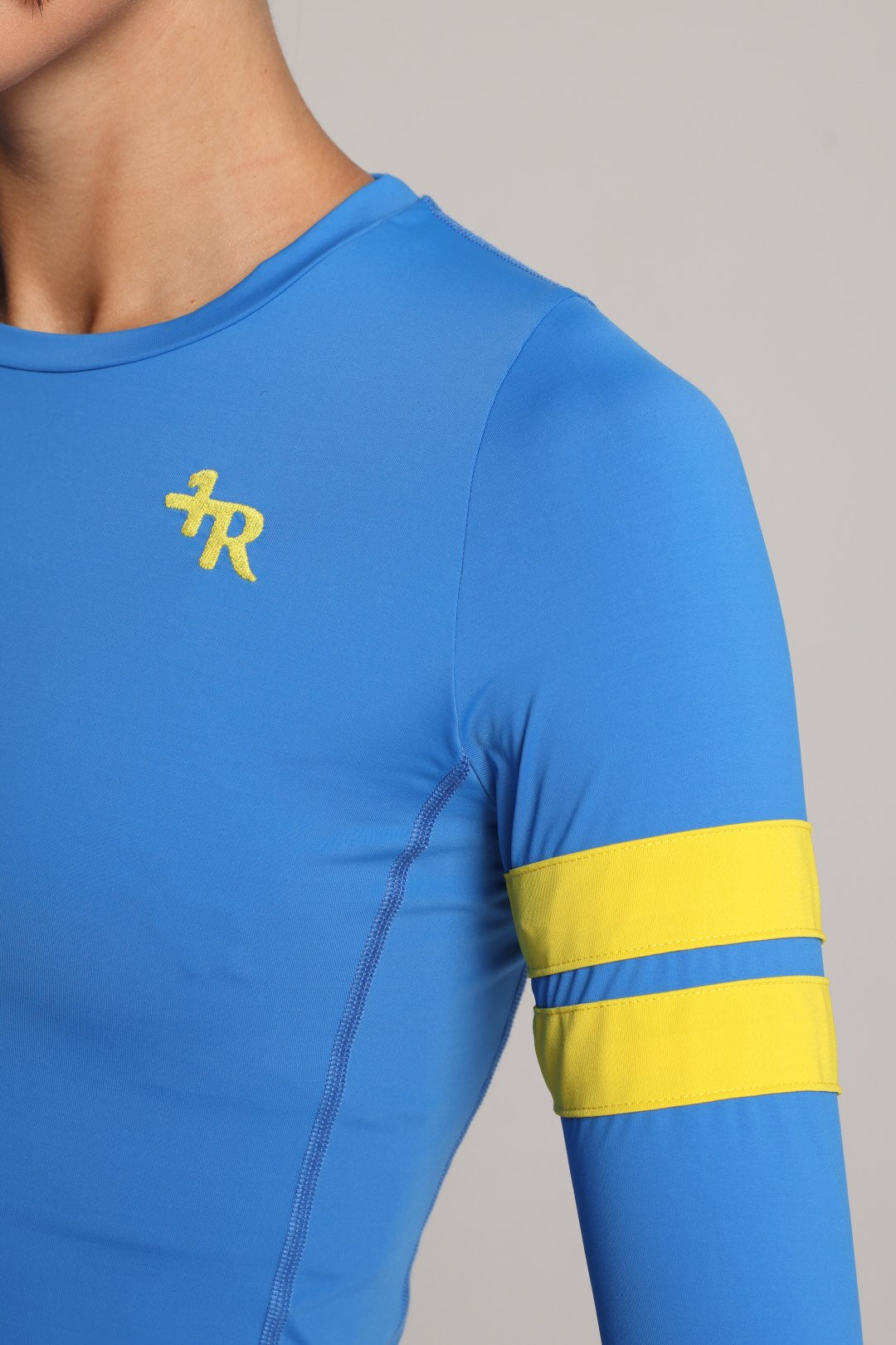 Close-up of a blue long-sleeve athletic top worn by a model, featuring a yellow stylized logo on the left chest and two horizontal yellow stripes on the right sleeve, against a light gray background - by One More Reason, the European premium sportswear brand.
