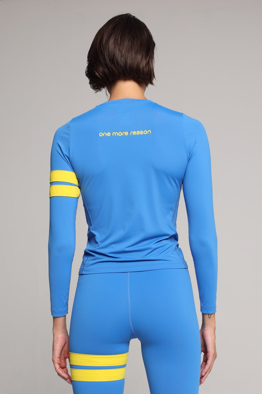 A woman shown from the back in a bright blue long-sleeve athletic top and matching leggings set with yellow stripe accents on the sleeves and thighs, against a plain light gray studio background. All by One More Reason, the European premium sportswear brand.