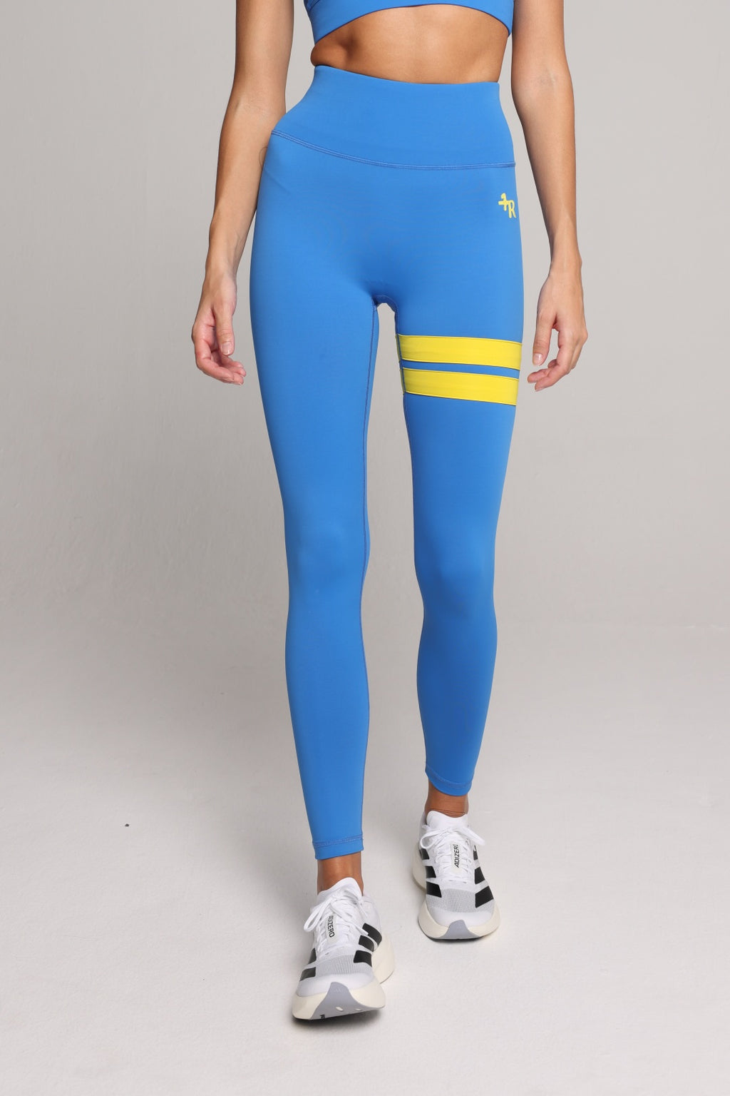A model wearing bright blue high-waisted leggings with two horizontal yellow stripes on the upper thigh and a small yellow logo on the hip, paired with white sneakers, against a light gray studio background. All by one more reason, the European premium sportswear brand 