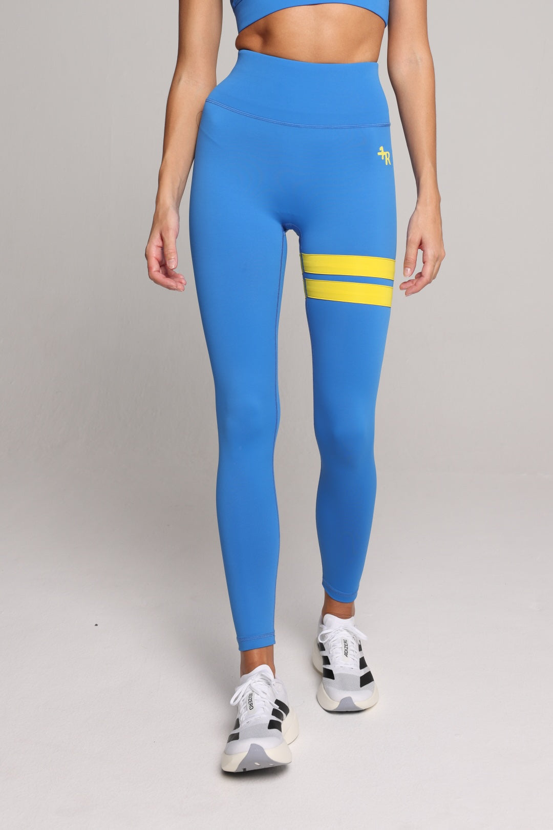 A model wearing bright blue high-waisted leggings with two horizontal yellow stripes on the upper thigh and a small yellow logo on the hip, paired with white sneakers, against a light gray studio background. All by one more reason, the European premium sportswear brand 