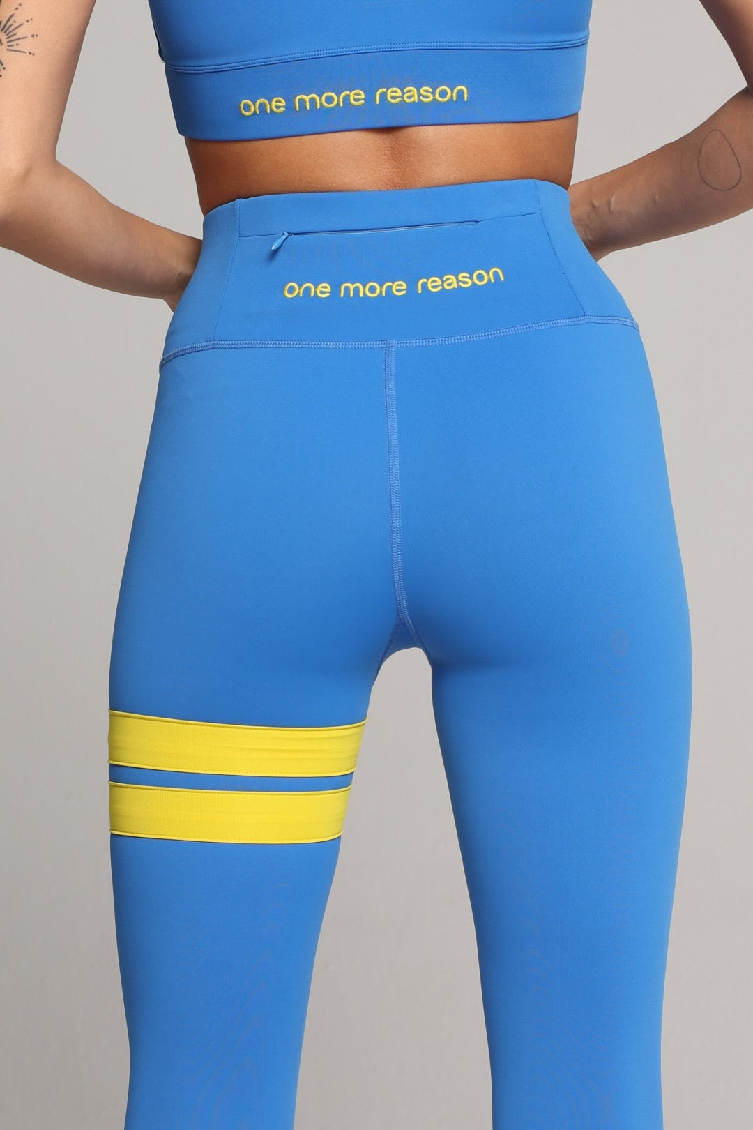 A woman shown from the back wearing bright blue high-waisted leggings with two horizontal yellow stripes on the upper thigh and a small yellow logo on the hip, paired with white sneakers, against a light gray studio background. All by one more reason, the European premium sportswear brand 