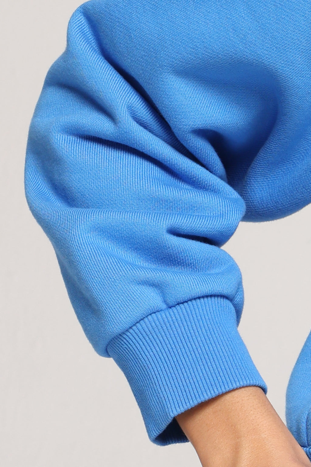 Close-up of a bright blue ribbed knit garment, likely a sweatshirt or sweater, showing the sleeve cuff and part of the arm against a light neutral background.