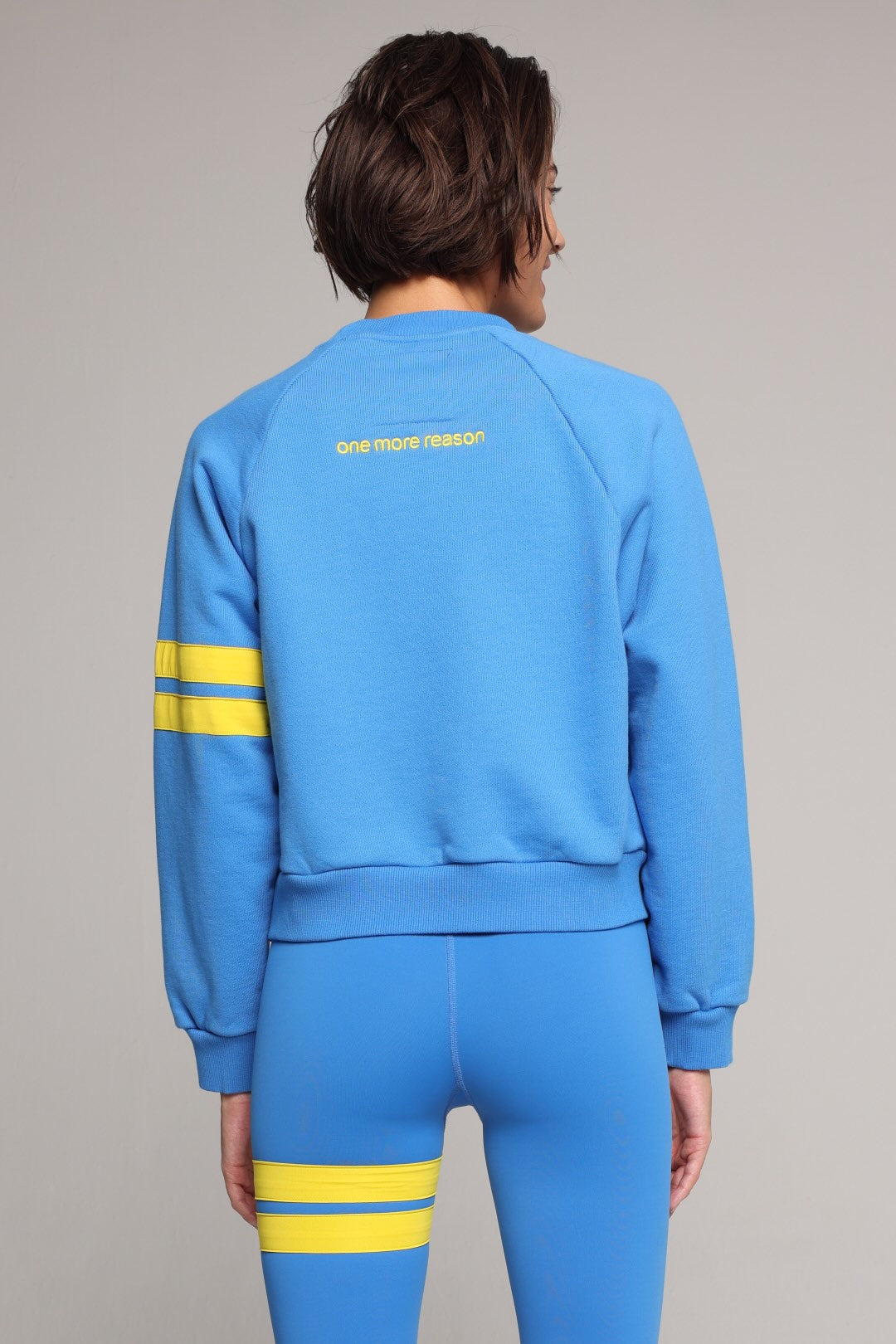 Back view of woman wearing blue high-waisted leggings and oversized  blue crew sweater with yellow stripes by One More Reason, studio shot.