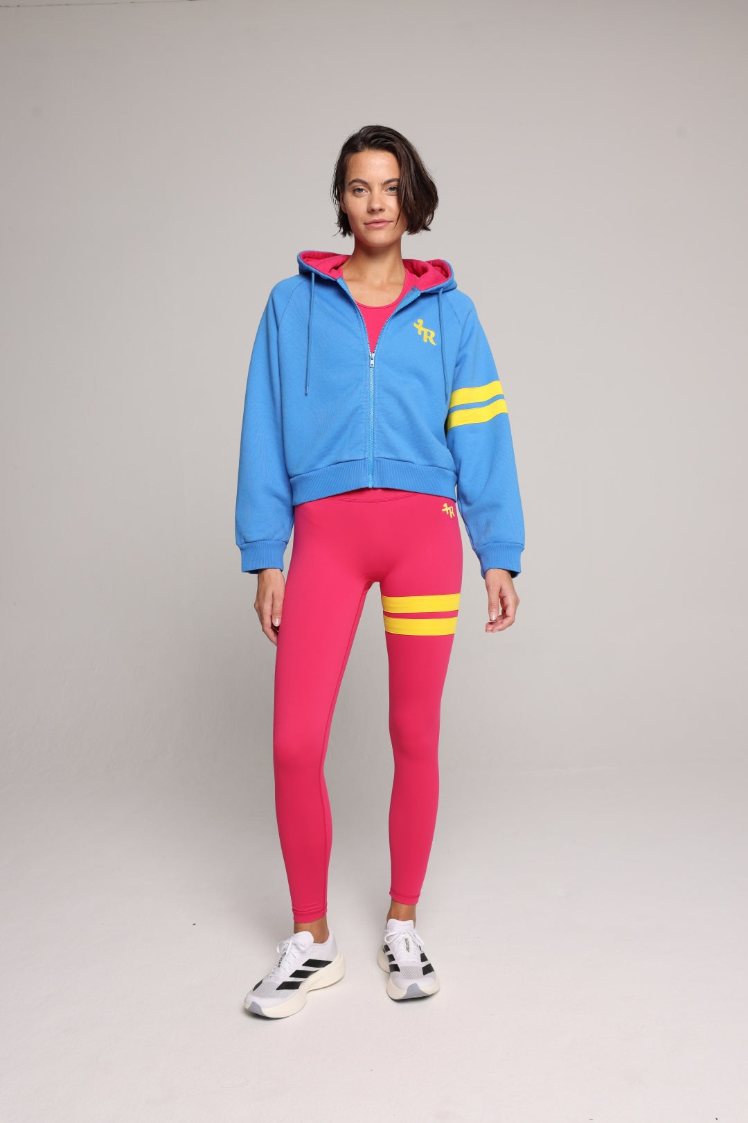 A woman wearing a blue zip hoodie and pink leggings with yellow stripes by One More Reason, the premium sportswear brand made in Europe in a studio setting.