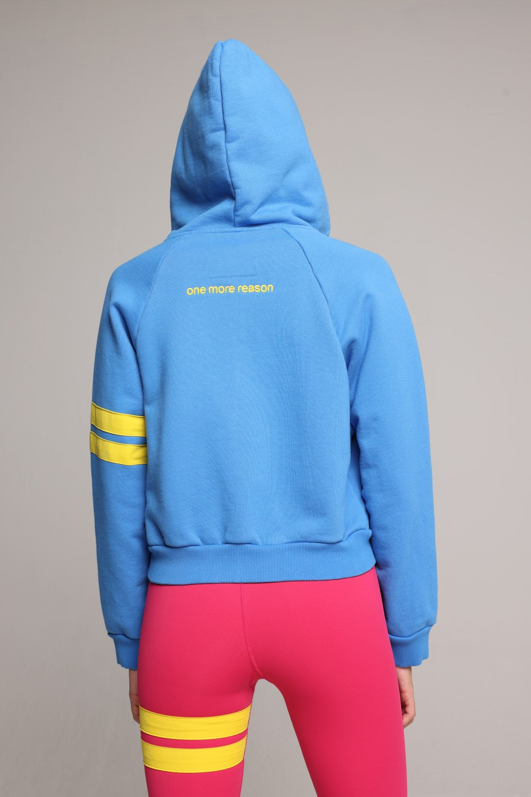 woman wearing blue and pink luxury athletic set, blue bicolor zip hoodie and pink tights by  One More Reason, with yellow stripes, studio look, back view