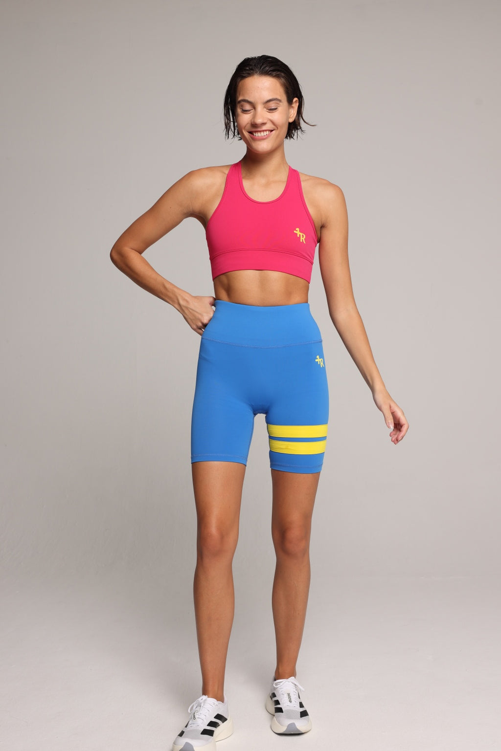 Woman wearing a soft pink sports bra and blue short leggings tights with yellow stripes by One More Reason fashion forward luxury sports wear, studio set