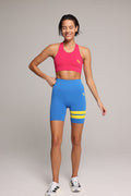 Woman wearing a soft pink sports bra and blue short leggings tights with yellow stripes by One More Reason fashion forward luxury sports wear, studio set