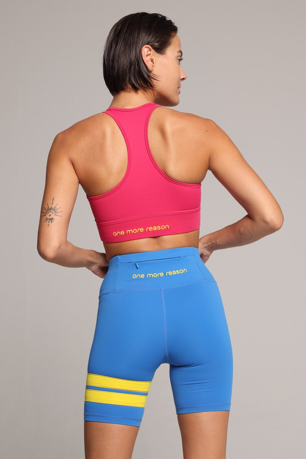 A woman shown from the back wearing a bright pink sports bra and blue fitted biker shorts with yellow stripes. The shorts have the words 'one more reason' printed in yellow on the back waistband. Premium Sportswear by One More Reason.