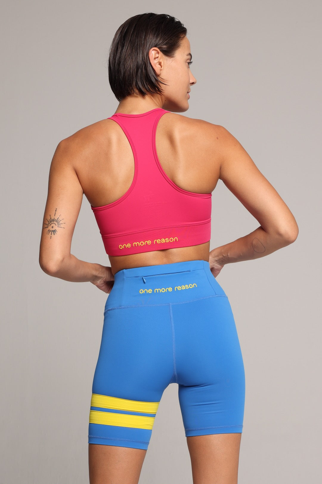 A woman shown from the back wearing a bright pink sports bra and blue fitted biker shorts with yellow stripes. The shorts have the words 'one more reason' printed in yellow on the back waistband. Premium Sportswear by One More Reason.