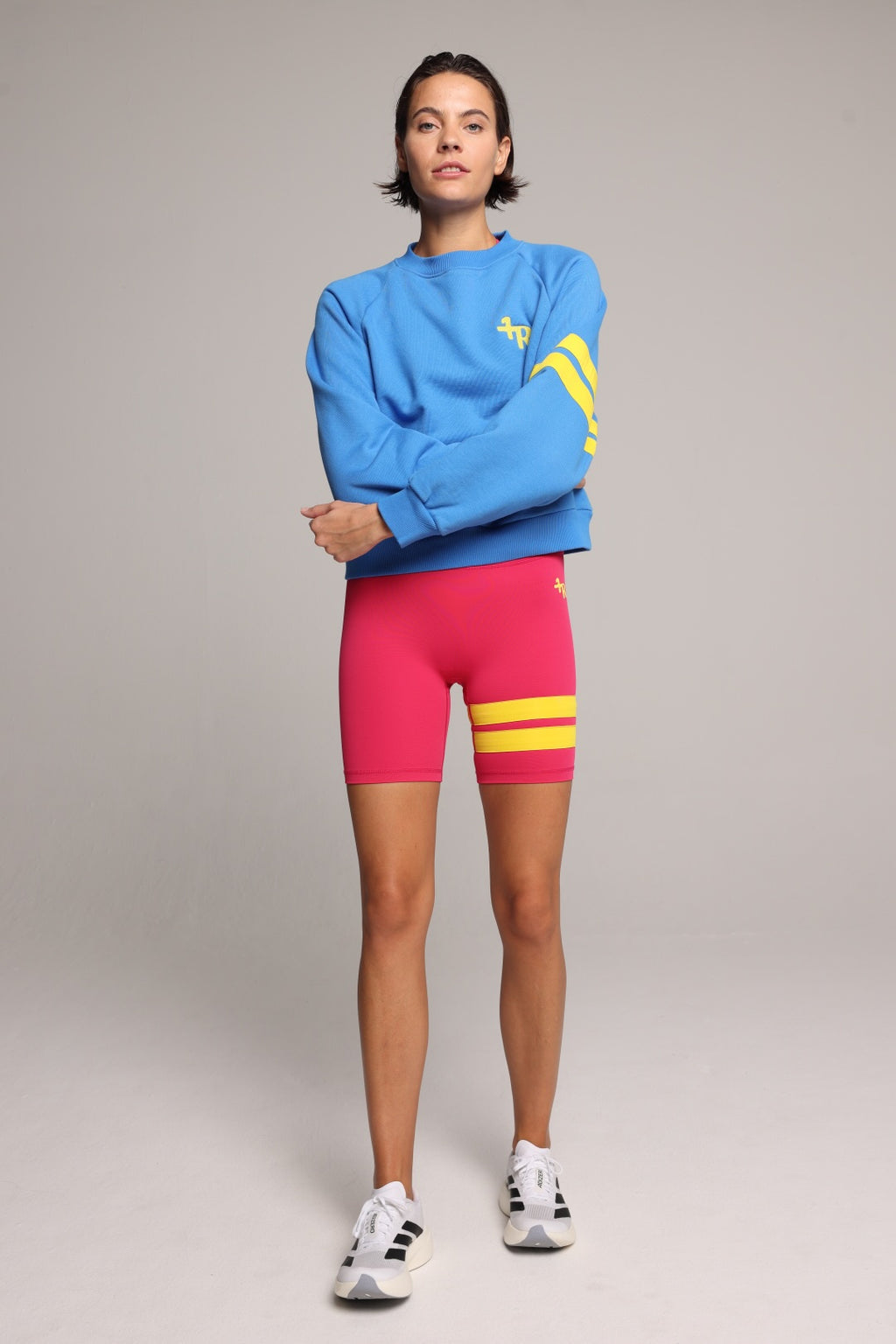 A model wearing a bright blue crewneck sweatshirt with yellow graphic stripes, hot pink biker shorts with yellow stripes, and white sneakers, standing against a neutral gray studio background. All by One More Reason, the premium European sportswear brand.