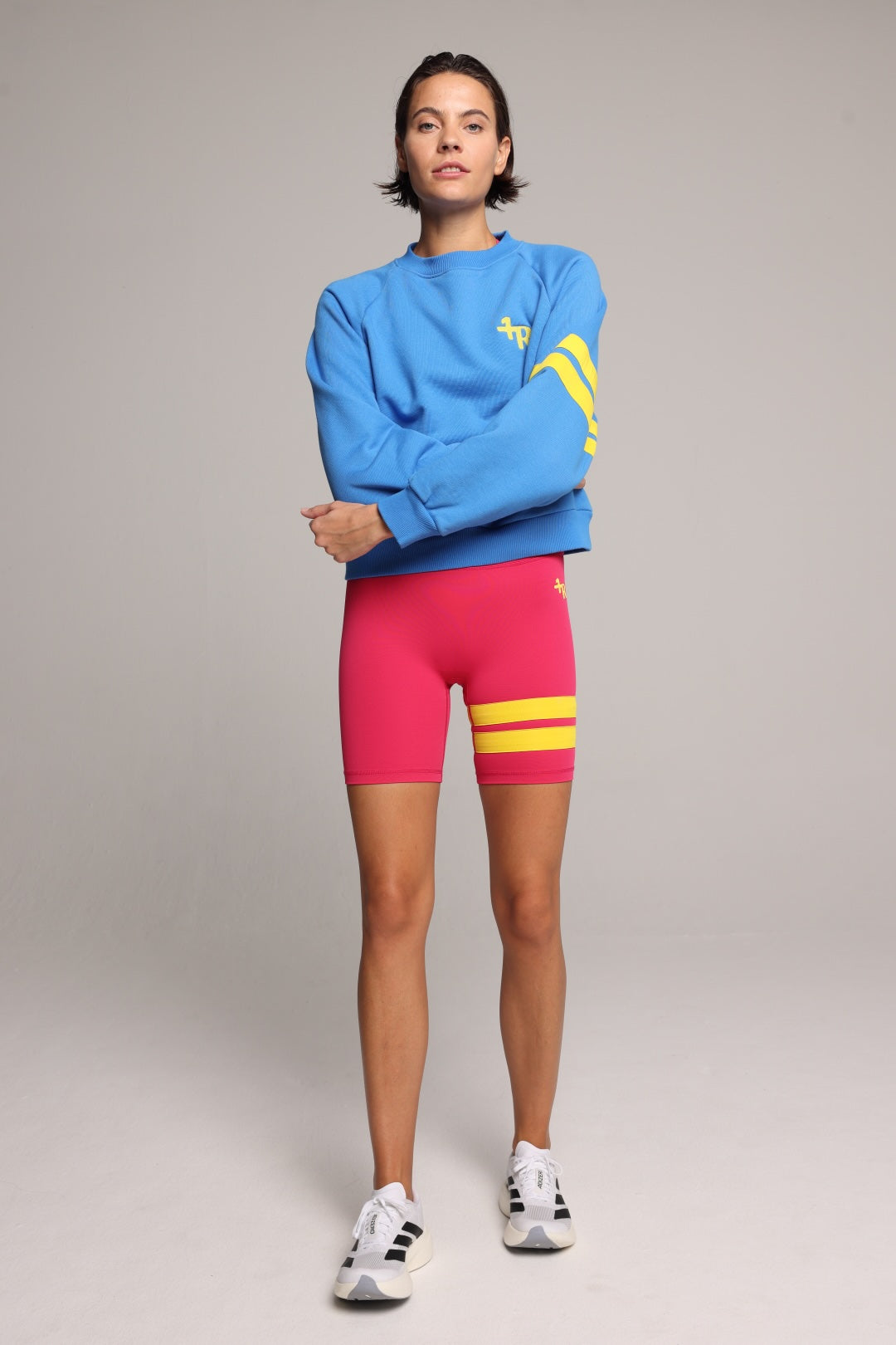 A model wearing a bright blue crewneck sweatshirt with yellow graphic stripes, hot pink biker shorts with yellow stripes, and white sneakers, standing against a neutral gray studio background. All by One More Reason, the premium European sportswear brand.