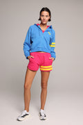 A woman in a bright blue zip-up hoodie with yellow stripes on the sleeve, hot pink shorts with yellow stripes on the hem, and white sneakers, against a neutral gray studio background. All by One More Reason, the premium European sportswear brand.