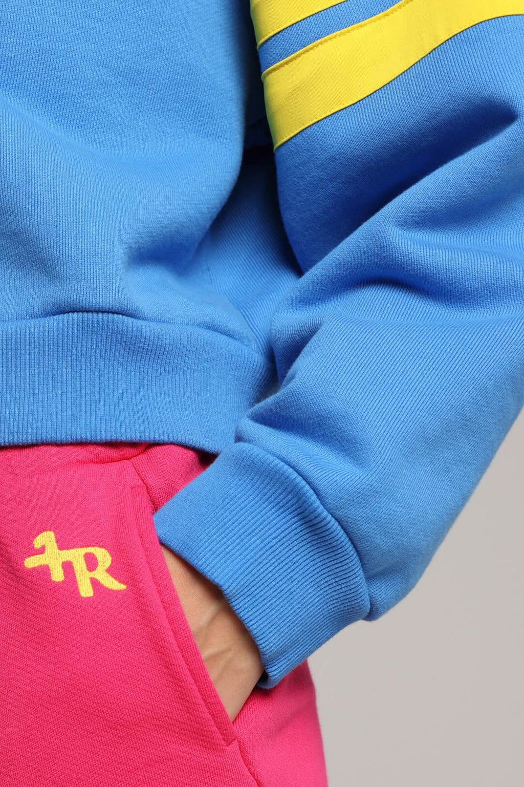 Close-up of a person wearing a bright blue sweatshirt with yellow stripes on the sleeve and a yellow monogram logo on pink pants. All from One More Reason, the European Premium Sportswear Brand.
