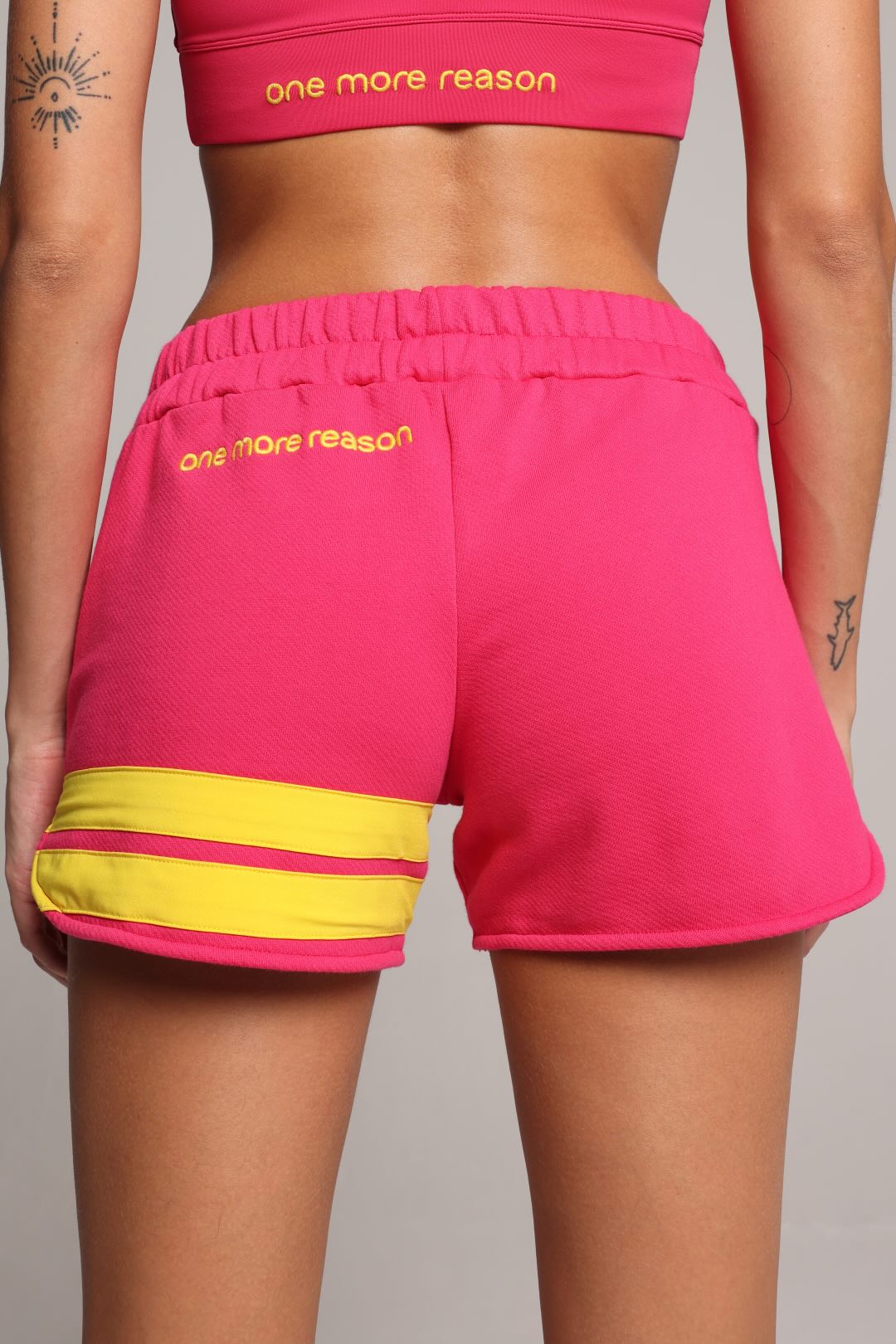 Bright pink athletic shorts with yellow stripe details at the hem and yellow 'one more reason' text on the back, shown on a model from the back.