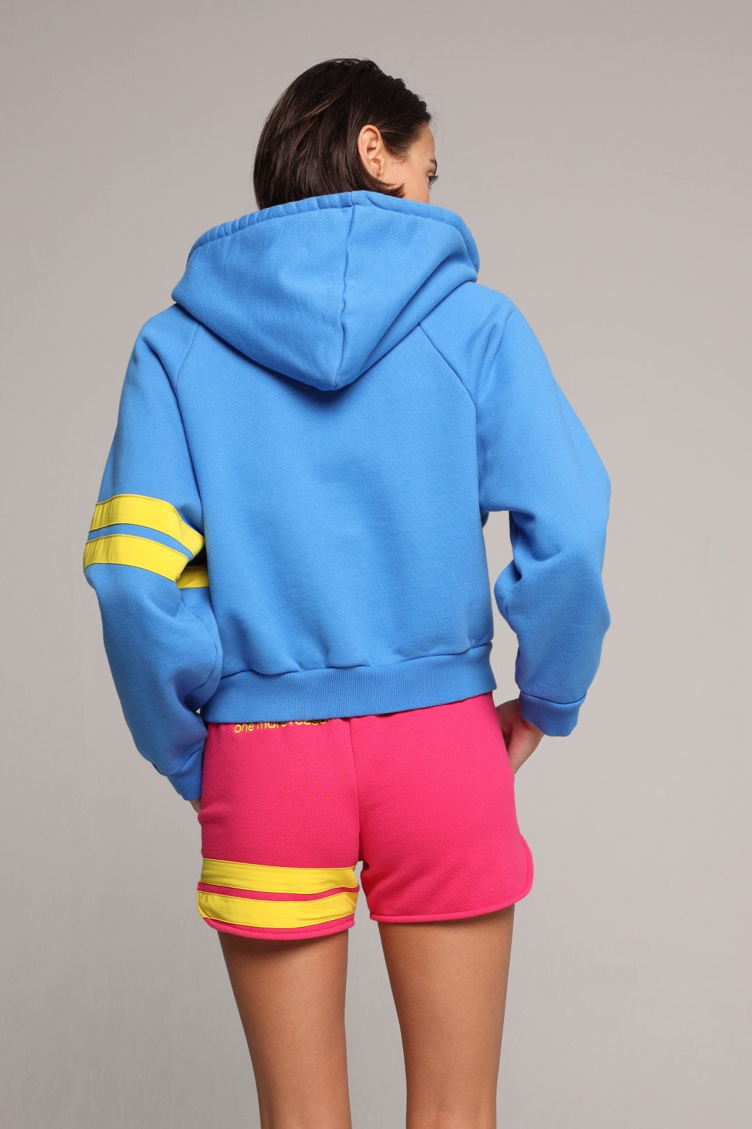 A woman showing from the back wearing a bright blue zip-up hoodie with a pink-lined hood, yellow stripes on the sleeves, and matching hot pink sweat shorts with yellow stripes on the hem. All from One More Reason, the European Premium Sportswear Brand.